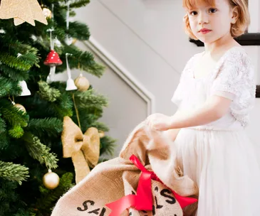 Young child with a Santa sack by a decorated Christmas tree with gold ornaments and red ribbon.