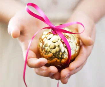 Hands holding a golden bauble with a pink ribbon, suggesting a festive decoration.