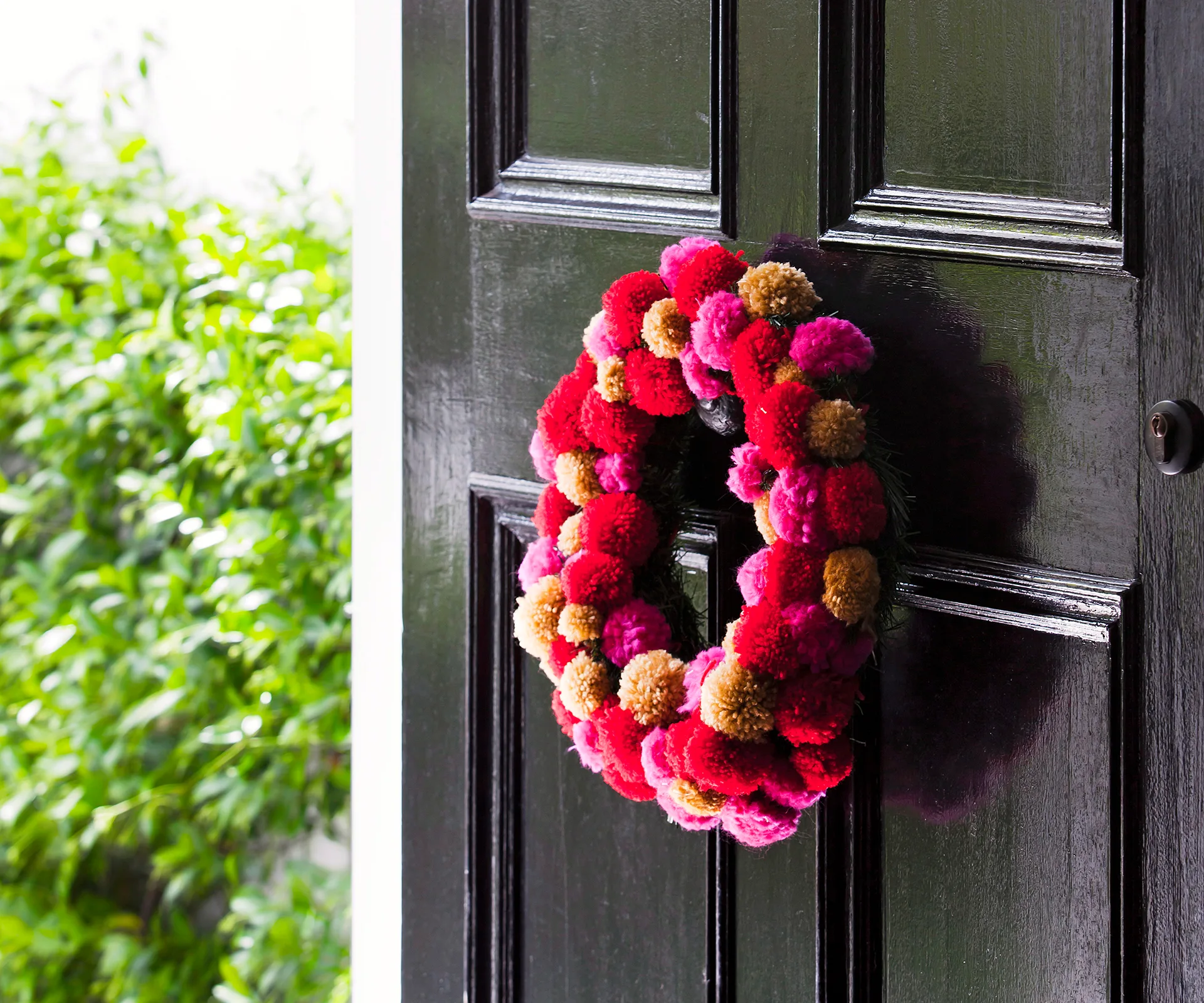 Colorful pompom wreath with red, pink, and yellow pom-poms on a dark door, greenery visible in the background.