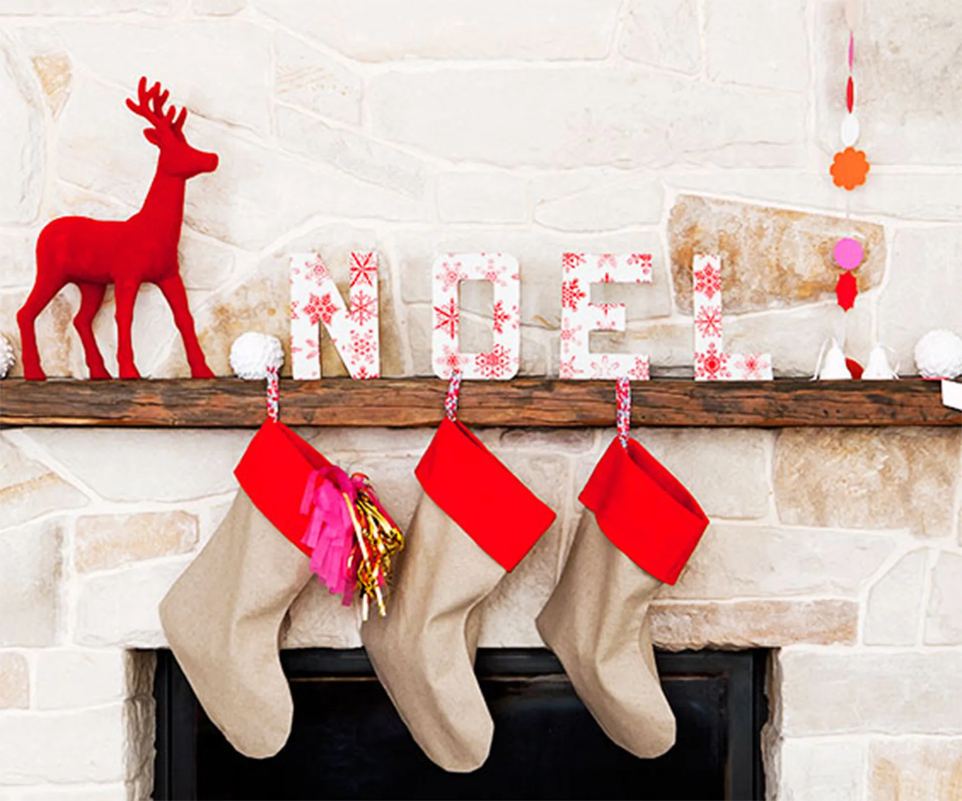 Red reindeer figurine and "NOEL" letters on a mantel with three hanging stockings against a stone background.