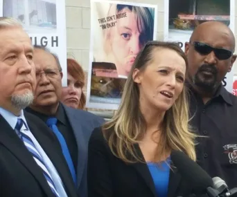 Group of people speaking at a press conference, woman in focus with a microphone. Posters in the background.
