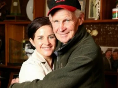 Father and daughter embrace warmly in a room with wooden shelves and photos in the background.