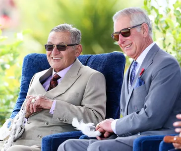 Two men in suits and sunglasses smile while seated outdoors on blue chairs.