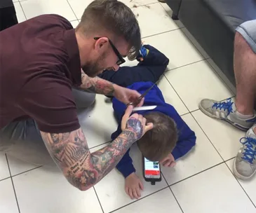Barber with tattoos cuts a young boy's hair on the floor while the boy watches a smartphone, providing a comfortable setting.