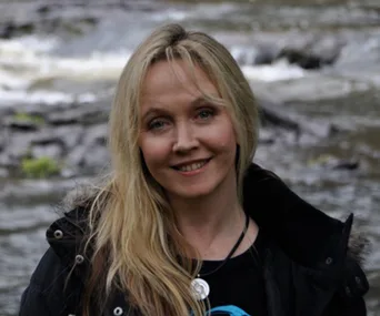A woman with long blond hair smiles outdoors with a river in the background, wearing a black jacket and necklace.