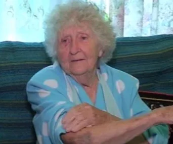 Elderly woman with curly white hair and blue robe sitting on a couch, appearing calm, with patterned curtains in the background.