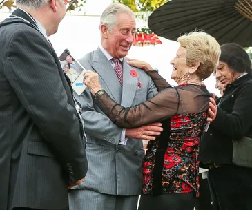 An elderly woman embraces and kisses a smiling older man in a suit, surrounded by onlookers.