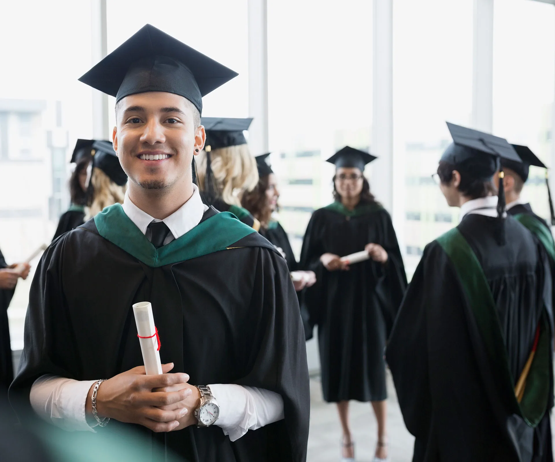 Graduates in caps and gowns gather with diplomas in hand, smiling and celebrating at a sunny indoor ceremony.