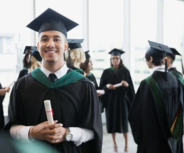 Graduates in caps and gowns gather with diplomas in hand, smiling and celebrating at a sunny indoor ceremony.