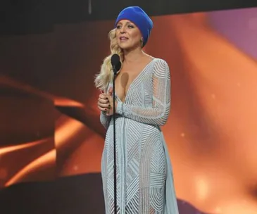 A woman in a blue beanie and patterned dress speaks at a microphone against an orange-lit background.