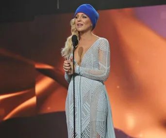 A woman in a blue beanie and patterned dress speaks at a microphone against an orange-lit background.