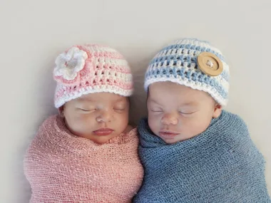 Twin babies sleeping, wrapped in pink and blue blankets, wearing matching crochet hats with flower and button details.