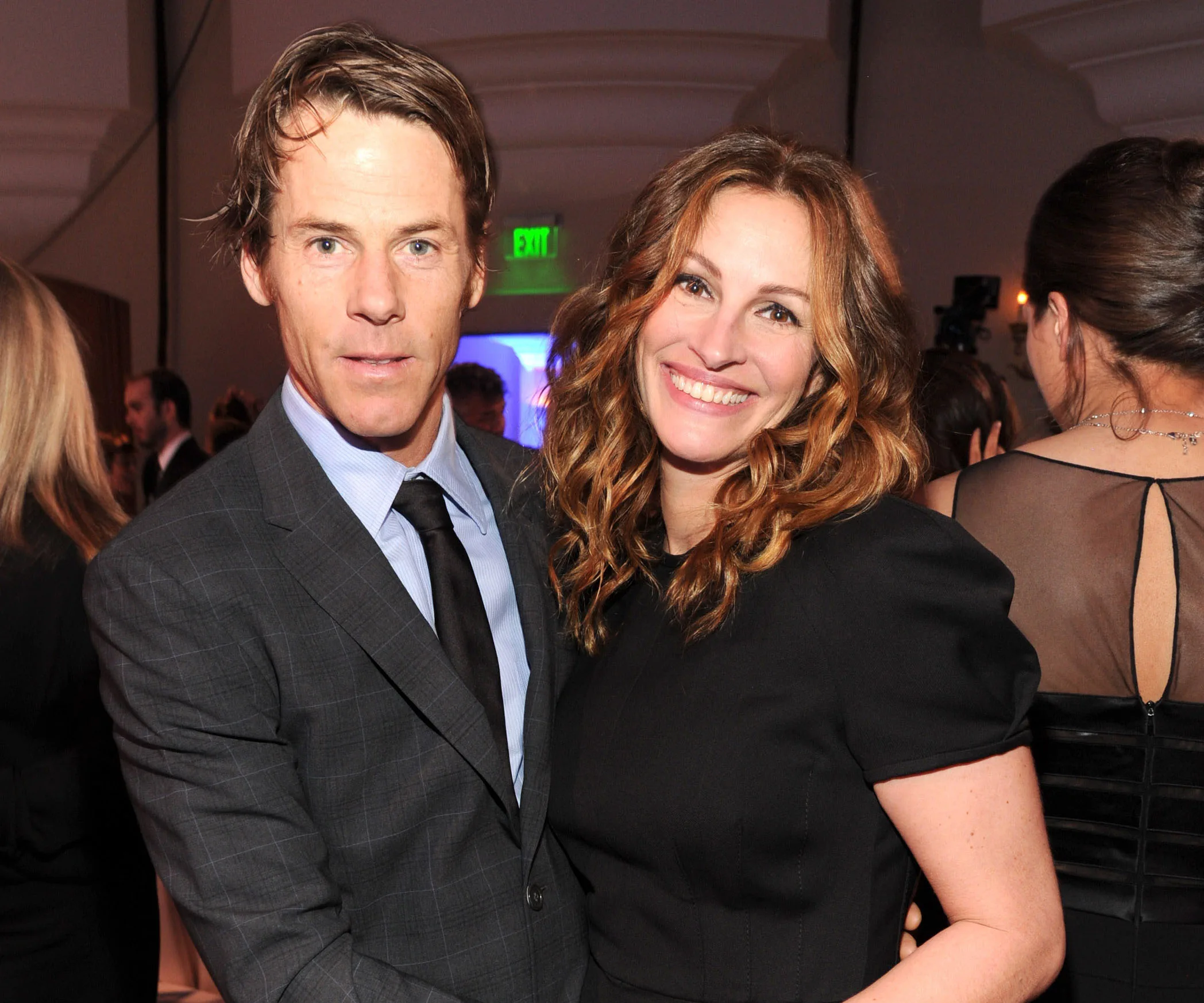 A smiling couple dressed elegantly at an indoor event, surrounded by other guests.