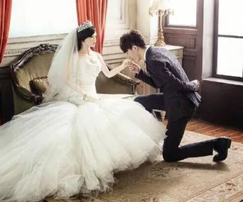 A groom kneels and kisses the hand of a life-sized doll in a wedding dress, seated on a vintage couch in a luxuriously decorated room.