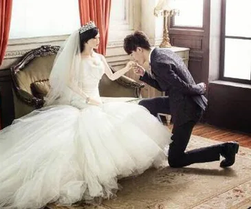A groom kneels and kisses the hand of a life-sized doll in a wedding dress, seated on a vintage couch in a luxuriously decorated room.