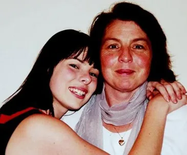 Two women smiling, one embracing the other, close-up shot against a plain background.