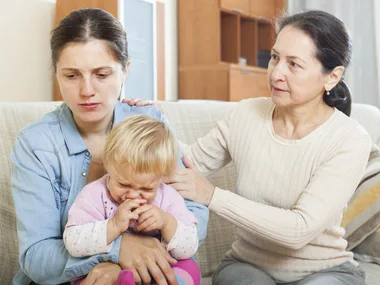 A concerned woman comforts a stressed mother holding a crying baby on a couch.