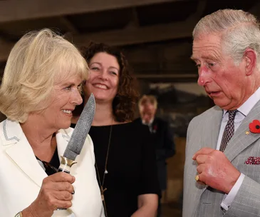Camilla smiling while holding a knife as Prince Charles looks on with surprise.