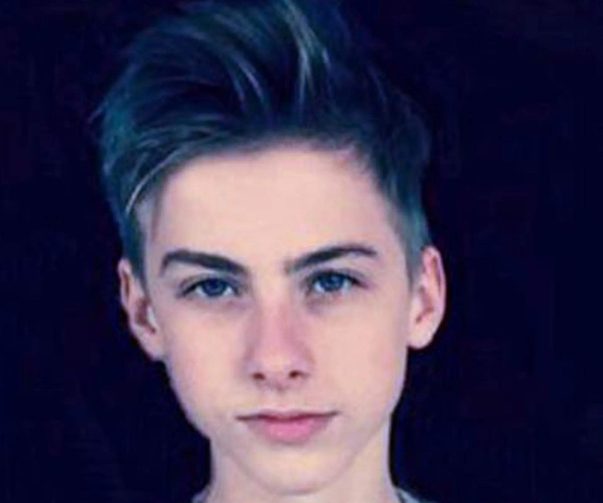 A young boy with short, styled hair and a serious expression against a dark background.