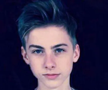 A young boy with short, styled hair and a serious expression against a dark background.