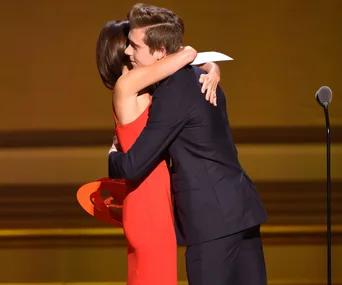 A woman in a red dress hugging a young man in a suit on stage, holding an award.