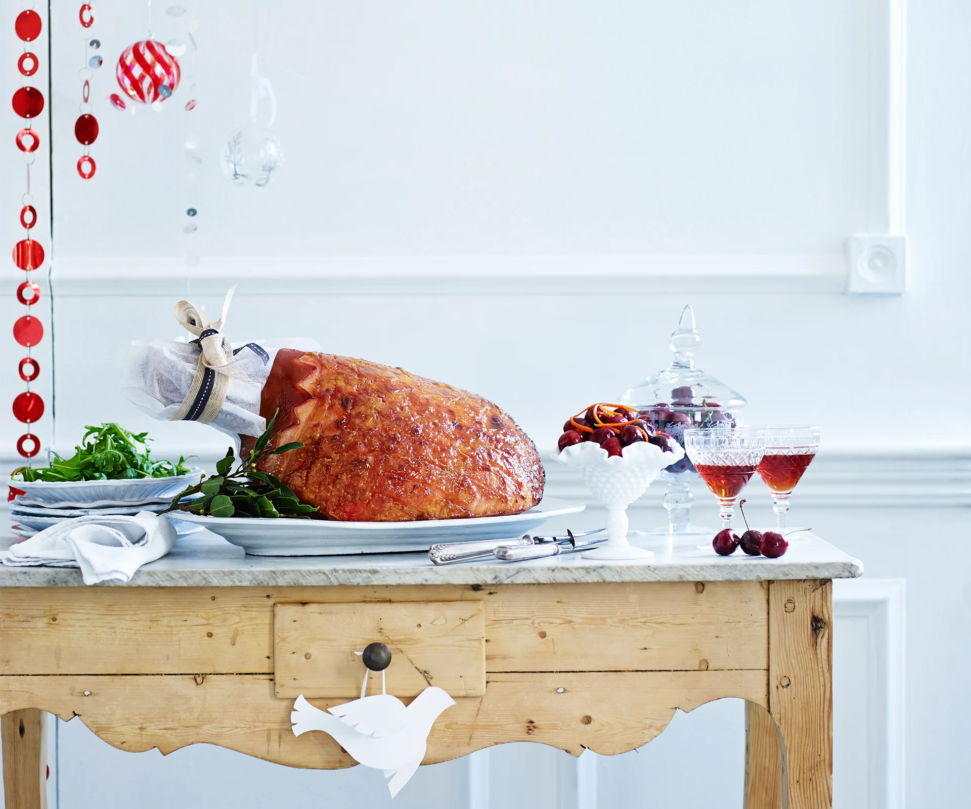 Roasted ham on a wooden table with salad, cherries, and drinks; festive decor in the background.