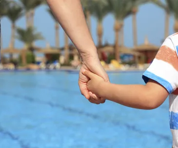 Adult and child holding hands near a pool with palm trees in the background.
