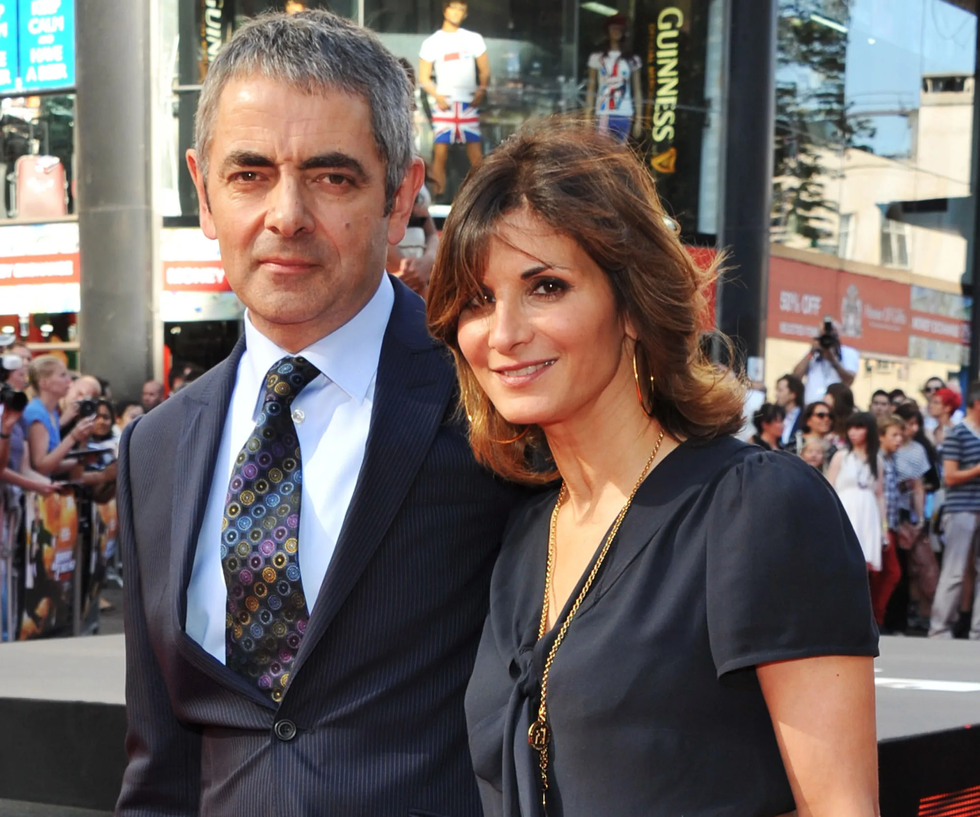 A man in a suit and a woman in a dark dress pose together at a public event with a crowd in the background.