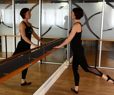 A woman practicing ballet exercises at a barre, reflected in a mirror, wearing a black tank top and leggings.