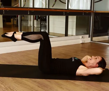 Person performing an ab exercise on a mat, legs raised, hands behind head in a fitness studio setting.