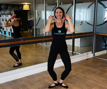 Person in a dance studio doing an exercise with small pink dumbbells, wearing an 'XTEND BARRE' tank top.