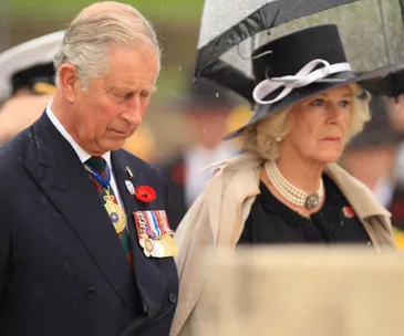 Royal ceremony attendees under rain, one in medals and the other in a hat and pearls, under an umbrella.
