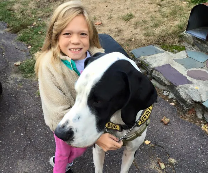 A young girl smiling while hugging a large Great Dane service dog on a paved path.