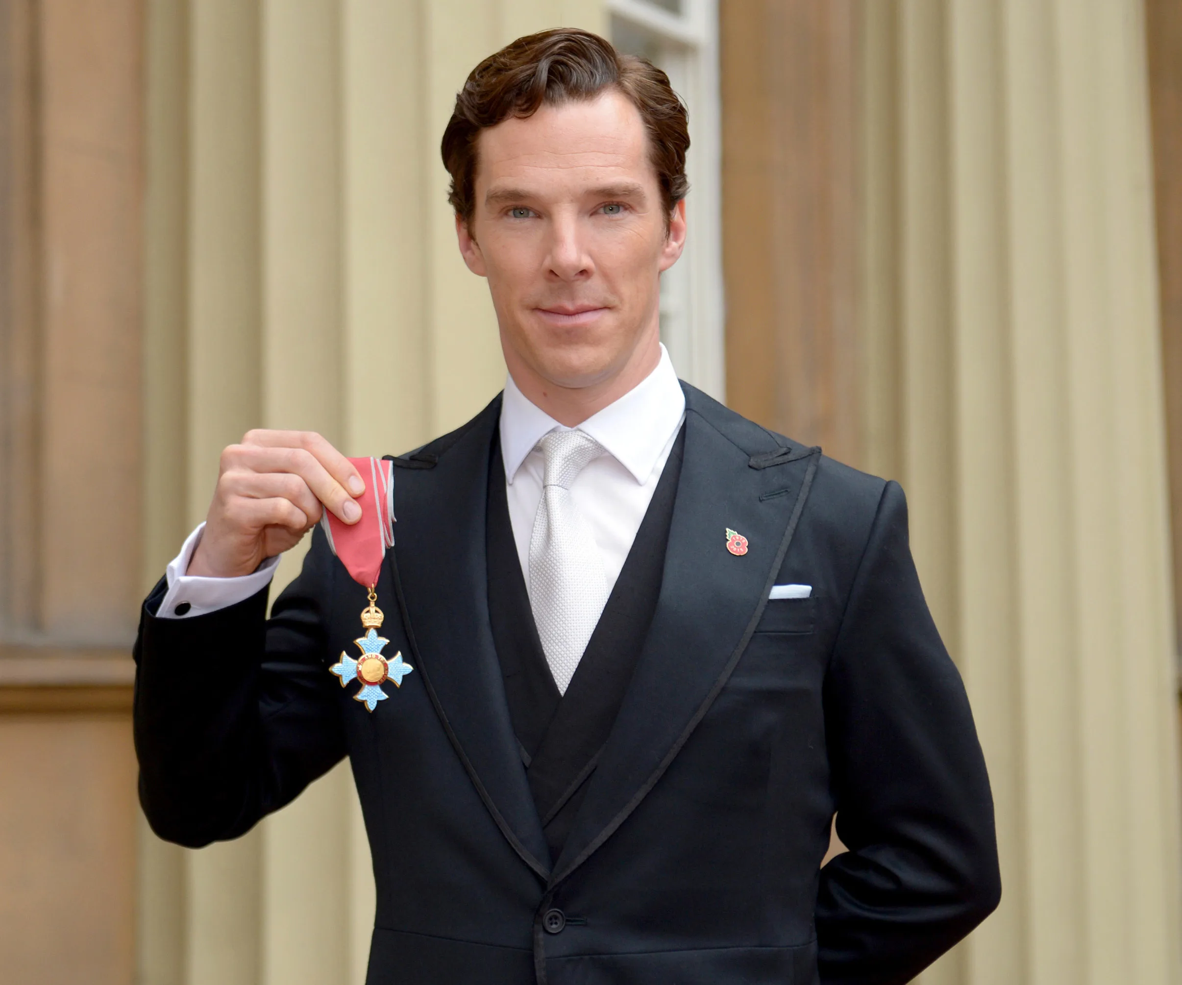 A person in a suit holds a CBE medal outside a grand building.
