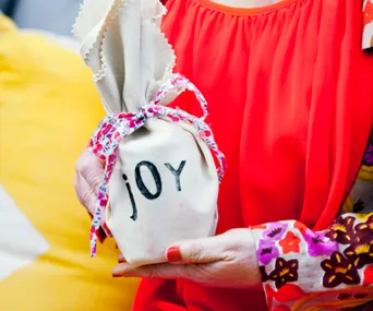 Person in red holding fabric-wrapped gift with "joy" text, tied with a floral ribbon.