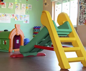 Colorful indoor playground with a small slide, plastic playhouse, and children's artwork on the walls.