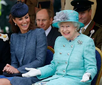 Two women in elegant hats and coats share a laugh, sitting at an event with seated military personnel in the background.