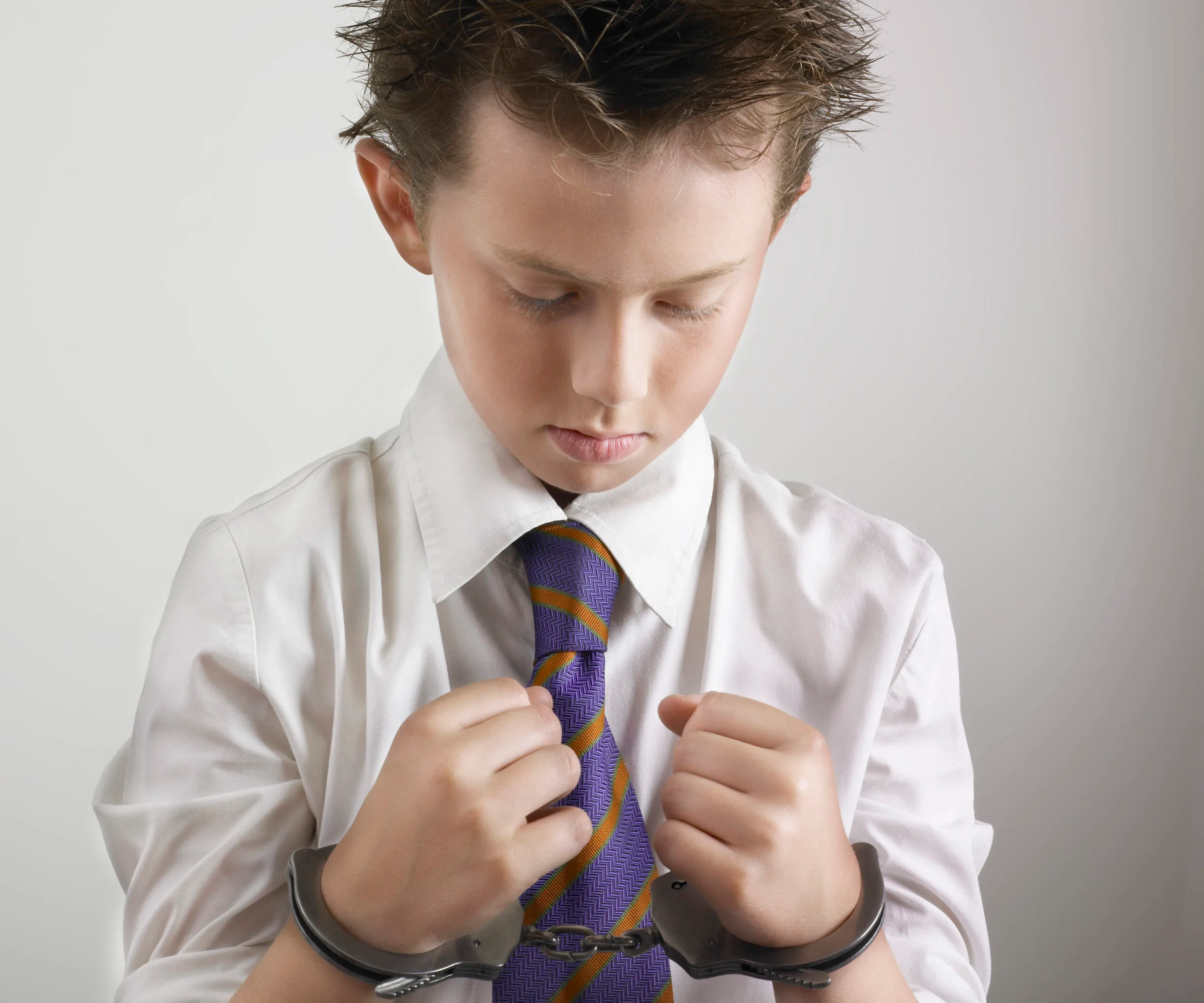 Young boy in handcuffs wearing a white shirt and striped tie, looking down with a serious expression.