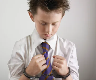 Young boy in handcuffs wearing a white shirt and striped tie, looking down with a serious expression.