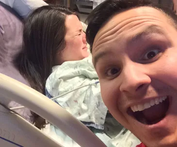 Smiling man takes selfie as woman behind him is in labor at hospital bed.