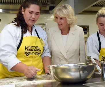 Royal visit to OzHarvest in Sydney; Camilla observes cooking activity with participants wearing yellow aprons.