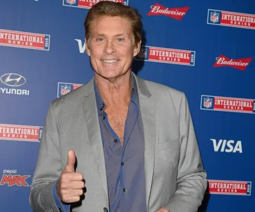 Man in a grey suit gives a thumbs up at an NFL event backdrop.