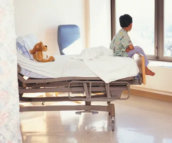 Child in hospital gown sitting on bed, looking out window, with a stuffed lion toy beside them.