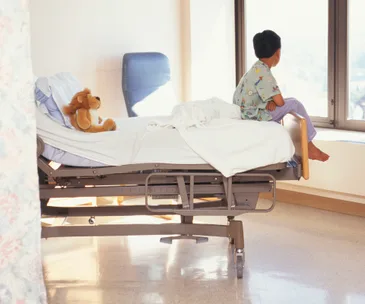 Child in hospital gown sitting on bed, looking out window, with a stuffed lion toy beside them.
