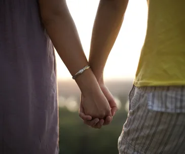 Two people holding hands at sunset, wearing casual summer clothing and bracelets.
