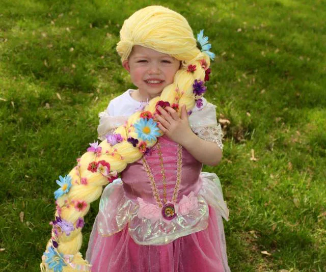 Child in a pink dress and blonde wig with flowers, resembling Rapunzel, smiling outdoors.