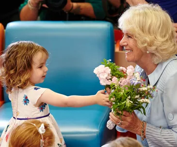 A smiling woman receives flowers from a young girl, both are seated with a blue chair in the background.