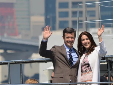 A man and a woman smiling and waving on a boat, with a cityscape in the background.