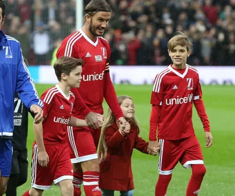 David Beckham and his children in red jerseys walking on a soccer field, smiling and holding hands.