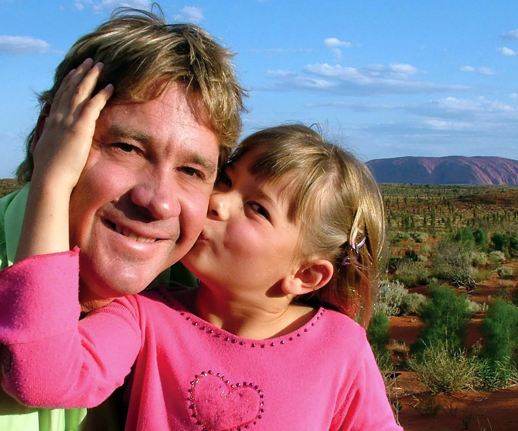 Father and young daughter share a sweet moment in a scenic outdoor setting with blue sky and landscape in the background.